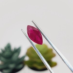 May include: A marquise-cut ruby gemstone held by tweezers. The ruby is a deep, vibrant red color, with facets catching the light. The tweezers are silver, and the background is blurred, with hints of green.