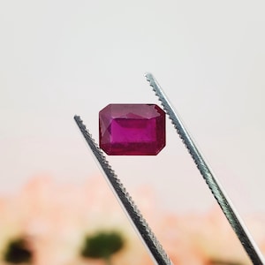 May include: A vibrant, deep red gemstone with a rectangular emerald cut, held securely by a pair of silver-toned metal tweezers. The stone features sharp, clean facets that reflect light, showcasing a rich, saturated crimson hue throughout its body.