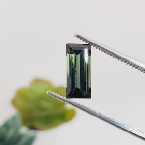 May include: A rectangular, emerald-green gemstone is held by a pair of silver tweezers. The gem has a dark, almost black, top and bottom, with vertical lines visible within the stone. The background is blurred, with a hint of green.