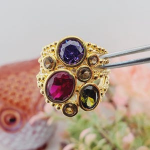 May include: A gold ring with multiple gemstones in various colors, including purple, red, and green. The ring has a textured band and is held by tweezers, showcasing the intricate design and craftsmanship of the jewelry.