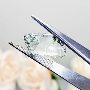 May include: A light green gemstone with a faceted cut is held by a pair of tweezers. The gemstone has a clear, translucent appearance with a slight green hue.