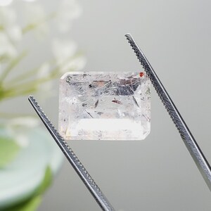 May include: A clear, rectangular gemstone with red and black inclusions, held by tweezers. The gemstone is transparent and has a slight shimmer.
