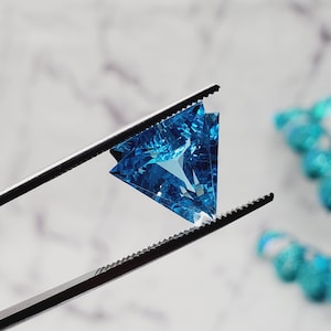 May include: A triangular blue topaz gemstone held by tweezers. The gemstone is faceted and has a brilliant shine.