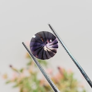 May include: A round, faceted gemstone held by tweezers. The gem features a swirl pattern of dark purple and white, with a polished surface. The tweezers are silver-colored, and the background is a soft, blurred white and green.