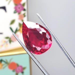 May include: A faceted, pear-shaped ruby gemstone held by silver tweezers. The ruby is a deep, vibrant red color and reflects light, showcasing its many facets. The tweezers are in sharp focus, highlighting the gemstone's clarity and brilliance.