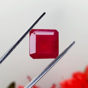 May include: A deep red gemstone with a square shape, held by a pair of tweezers.