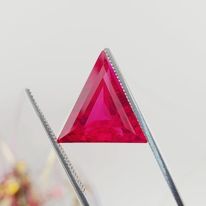May include: A triangular, faceted ruby gemstone with a deep red color. The gemstone is held in place by a pair of tweezers.