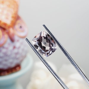 May include: A pale pink gemstone with a cushion cut, held by tweezers. The gemstone is clear and has a slight shimmer.