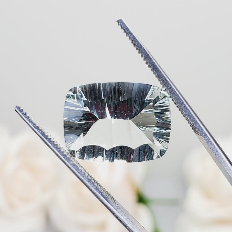 May include: A light green gemstone with a rectangular shape, held by tweezers. The gemstone has a faceted cut and a clear, translucent appearance.