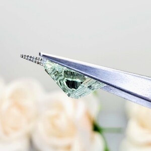 May include: A light green gemstone with a faceted cut, held by tweezers. The gemstone is clear and has a slight shimmer.