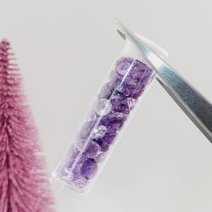 May include: A clear glass tube filled with small, purple and clear gemstones. The tube is held by a pair of tweezers.