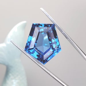May include: A faceted, pentagon-shaped gemstone in shades of blue, held by a pair of tweezers. The gem has a complex internal structure, reflecting light. The background is a soft, neutral color, highlighting the gem's brilliance. The gem is likely for jewelry making.