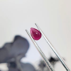May include: A small, teardrop-shaped, deep pink gemstone held securely between the serrated metal tips of a pair of silver-toned tweezers. The stone features a smooth, polished surface with a subtle sheen, reflecting light on its rounded top.