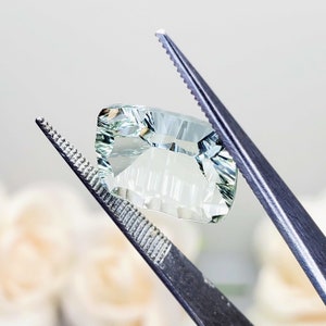 May include: A light green gemstone with a faceted cut, held by tweezers. The gemstone is clear and has a slight yellow tint.