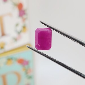 May include: A vibrant, rectangular-cut, magenta gemstone is held by a pair of silver tweezers. The gem has a polished surface and is set against a soft, blurred background. The gem's color is a rich, saturated pink.