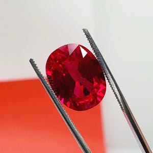 May include: A faceted, oval-shaped, deep red gemstone is held by a pair of silver tweezers. The gem reflects light, showcasing its internal structure and brilliance. The background is a gradient of white and red.