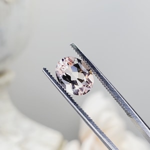 May include: A pale pink oval-shaped gemstone held by tweezers. The gemstone has a faceted cut and is translucent.