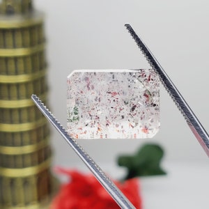 May include: A clear, faceted gemstone with red, green, and brown inclusions. The gemstone is held by tweezers against a white background.