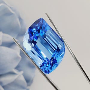 May include: A large, rectangular, blue topaz gemstone with facets. The gemstone is held by tweezers.