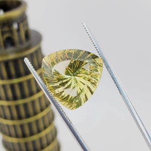 May include: A triangular, faceted gemstone with a yellow and green color pattern. The gemstone is held in tweezers.