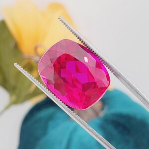 May include: A vibrant, cushion-cut gemstone with a deep magenta-pink hue and multifaceted surface, held securely by a pair of silver-toned metal tweezers. The stone displays high clarity and internal light reflection across its polished facets.