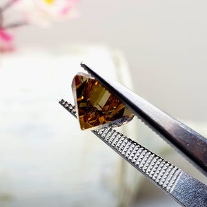 Puede incluir: Una piedra preciosa de color marr&oacute;n y dorado con forma triangular est&aacute; sujeta por unas pinzas. La piedra preciosa tiene un corte facetado y es transl&uacute;cida.