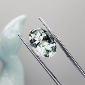 May include: A light green oval gemstone with a star-like pattern is held in place by a pair of silver tweezers.