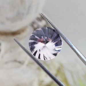 May include: A faceted, round gemstone with a swirling pattern of light and dark gray facets. The gem is held by a pair of silver tweezers, showcasing its intricate design and reflective qualities. The gem is a beautiful example of fine jewelry.