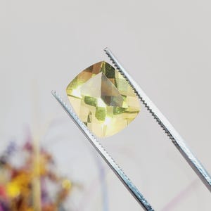 May include: A faceted, square-cut gemstone held by silver tweezers. The gem is a pale yellow color with multiple facets that reflect light. The background is a soft, blurred white.