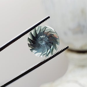 May include: A close-up of a green and white gemstone with a swirling pattern, held by tweezers.