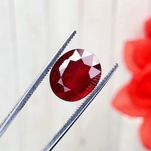 May include: A faceted, oval-shaped ruby gemstone held by a pair of silver tweezers. The ruby is a deep, rich red color with multiple facets that reflect light. A red flower is blurred in the background.
