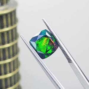 Puede incluir: Una piedra preciosa suelta de corte cojín sostenida por pinzas de metal plateado. La piedra presenta un color base oscuro con destellos iridiscentes y vibrantes de verde neón, amarillo y naranja en toda su superficie facetada, creando color.