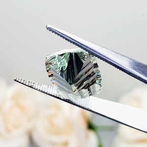 May include: A light green gemstone with a faceted cut, held by tweezers. The gemstone has a unique, striated pattern.