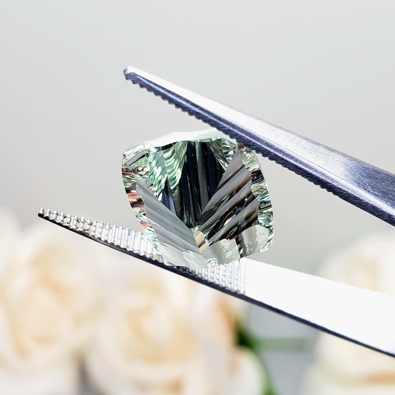 May include: A light green gemstone with a faceted cut, held by tweezers. The gemstone has a unique, striated pattern.