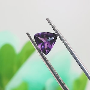May include: A triangular, faceted, deep purple gemstone is held by a pair of silver tweezers. The gem has a polished surface with internal facets that reflect light. The background is a soft, blurred mix of green and white.