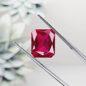 May include: A deep red gemstone with a square cut, held by tweezers against a white background.