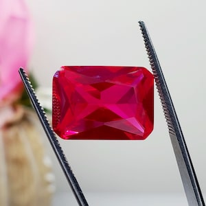May include: A vibrant, rectangular-cut, ruby-red gemstone is held by a pair of tweezers. The gem's facets reflect light, showcasing its clarity and brilliance. The background is softly blurred, highlighting the gem's color and shape.