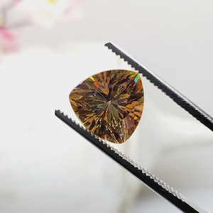Puede incluir: Una piedra preciosa tallada en forma triangular con un color marr&oacute;n dorado y facetas iridiscentes. La piedra preciosa est&aacute; sujeta por unas pinzas.