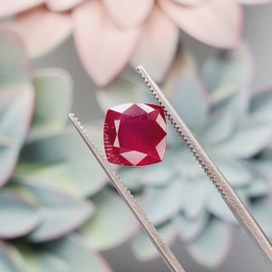 May include: A deep red gemstone with a cushion cut, held by tweezers against a blurred background.