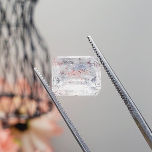 May include: A clear, faceted gemstone with pink inclusions, held by tweezers. The gemstone is rectangular in shape and has a slightly cloudy appearance.