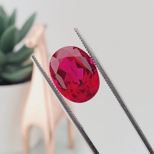 May include: A vibrant, oval-cut gemstone with a deep, saturated red color and multifaceted surface, held securely by a pair of silver-toned metal tweezers. The stone displays clear internal reflections and a polished, brilliant finish throughout.