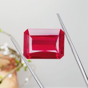 May include: A vibrant red gemstone with a rectangular emerald cut, held by tweezers against a white background.
