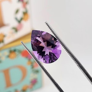 May include: A pear-shaped, faceted amethyst gemstone with a deep purple hue held by a pair of silver-toned metal tweezers. The gemstone displays a clear, brilliant cut with multiple triangular facets that reflect light across its surface.