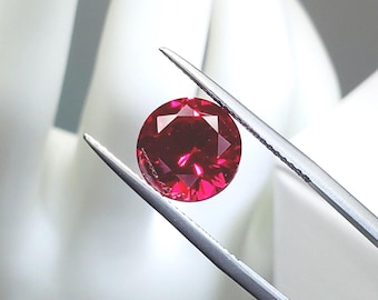 May include: A faceted, round, deep red gemstone is held by silver tweezers against a white background. The gem's cut allows light to reflect, creating bright highlights. This gemstone is likely for jewelry making.