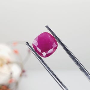 May include: A cushion-cut, vibrant pinkish-red gemstone held by a pair of serrated metal tweezers. The gemstone features a faceted surface that reflects light, showcasing a deep, saturated color and a symmetrical, rounded square shape.