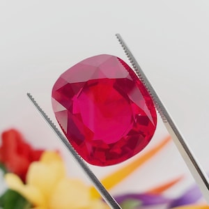 May include: A vibrant red gemstone with a cushion cut, held by tweezers against a blurred background of colorful flowers.
