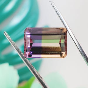 May include: A rectangular, emerald-cut gemstone featuring a distinct color gradient. The left side displays shades of purple and violet, transitioning into horizontal bands of green, yellow, and orange on the right side. The stone is held by metal tweezers.