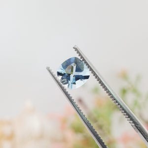 Puede incluir: Una piedra preciosa de color azul claro con facetas en talla cojín, sostenida por unas pinzas de metal plateado. La piedra tiene un aspecto claro y transparente con facetas geométricas que reflejan la luz, creando un tono azul frío y brillante.