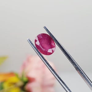 May include: A vibrant red oval-shaped gemstone held by a pair of tweezers. The gemstone has a slightly sparkly appearance.