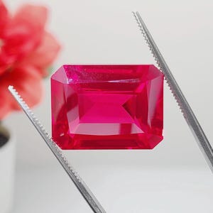 May include: A vibrant pink gemstone with a rectangular emerald cut, held by tweezers against a white background.